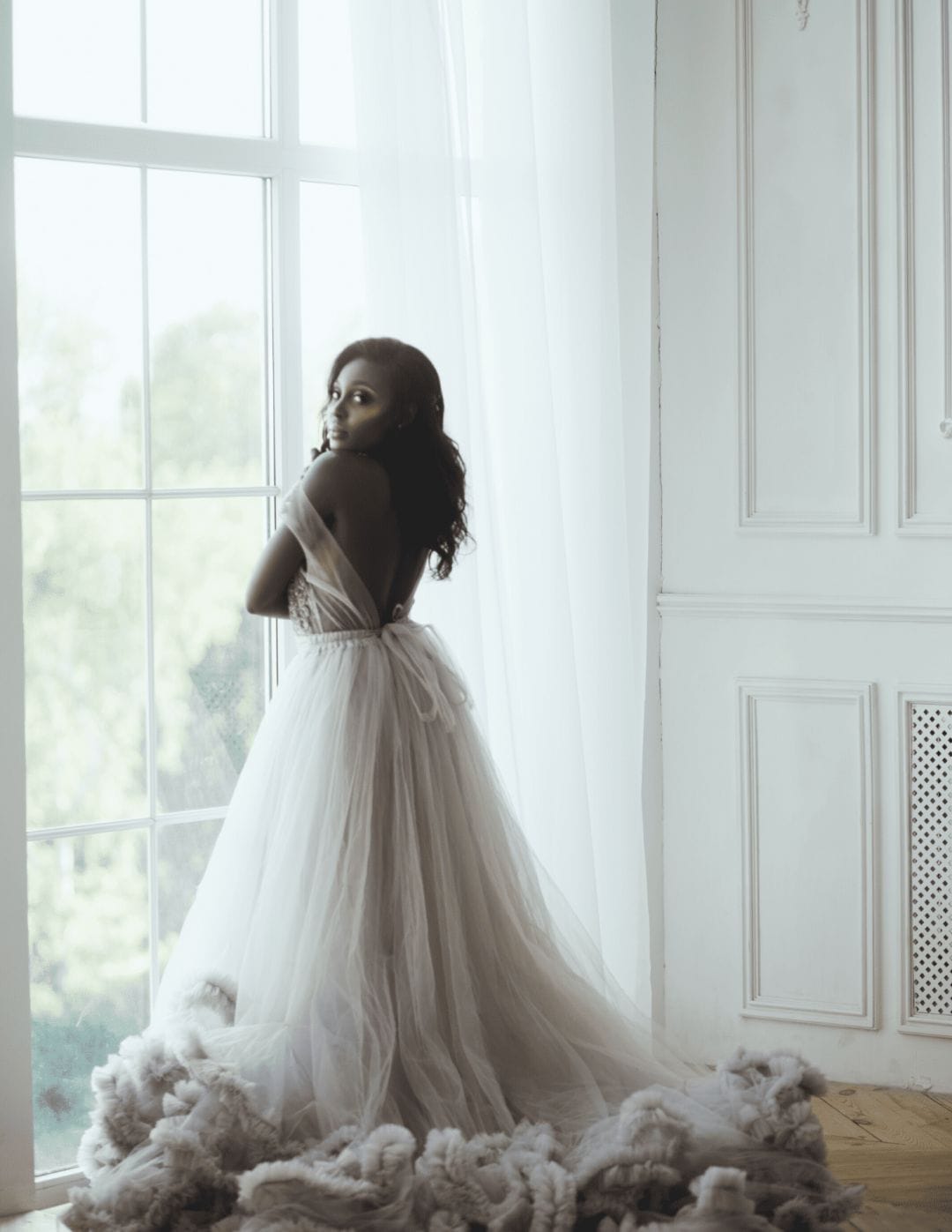 An African-American bride in her couture wedding dress looking over her shoulder while standing in front of a floor to ceiling window. The dress has lots of flowing tulle and ruffles.